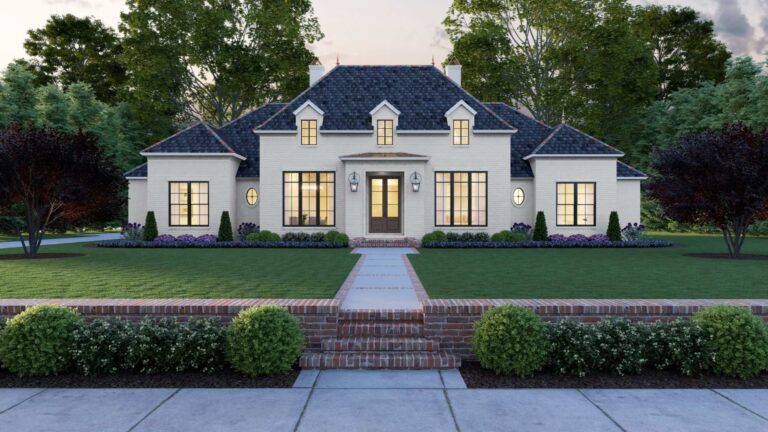 A front view of a French-inspired home with a sophisticated and timeless design. The exterior is finished with light-colored brick, complemented by a steeply pitched dark shingled roof. The house features a symmetrical layout with a central entrance framed by large windows on either side. The front door is flanked by lantern-style lights and leads up a brick staircase from a landscaped front yard. Above the entrance, two dormer windows add architectural interest to the roofline. The yard is well-manicured with a brick border, bushes, and flowering plants, set against a backdrop of mature trees and a soft, evening sky.