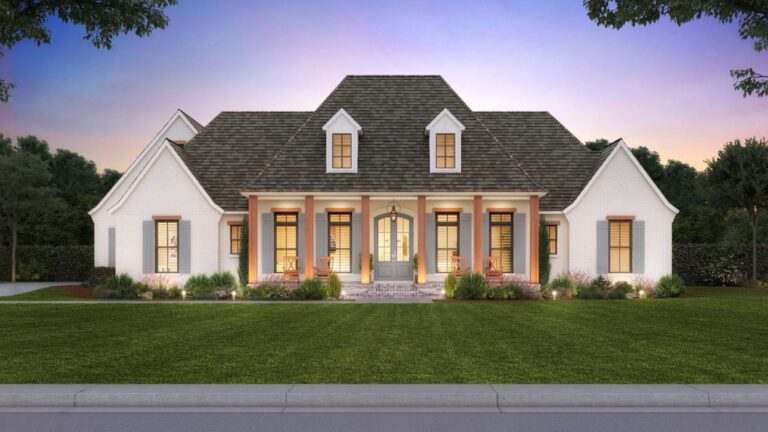 A front view of a traditional Southern-style home with a stately and elegant design. The exterior is finished with light-colored brick and a dark shingled roof. The home features a large, covered front porch supported by natural wood columns, leading to a central arched front door with glass panels. Flanking the entrance, large windows with shutters provide symmetry and balance. Above the porch, two dormer windows add architectural interest to the roofline. The front yard is well-manicured with a lush green lawn, shrubs, and flower beds, set against a backdrop of mature trees and a sky glowing with the colors of sunset.