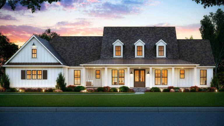 A front view of a modern farmhouse-style home with a warm and inviting design. The exterior features white board and batten siding, a dark shingled roof, and a metal roof over the large, covered front porch. The porch is supported by white columns and includes a front door with glass panels and a porch swing. Above the porch, there are three dormer windows that add character to the roofline. The windows are framed with dark shutters, complementing the overall color scheme. The front yard is landscaped with a lush green lawn, shrubs, and colorful flower beds, with trees and a sky painted with the colors of sunset in the background.