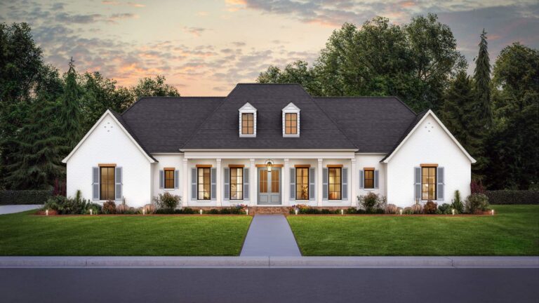 A front view of a traditional, elegant home with a symmetrical design and classic architecture. The exterior is finished with white brick, contrasted by a dark shingled roof. The central entryway features a covered front porch supported by white columns, leading to double front doors with glass panels. The house has large windows with shutters, and two dormer windows above the porch, adding character to the roofline. The front yard is well-maintained with a lush green lawn, shrubs, and neatly arranged flower beds. The home is set against a backdrop of tall trees and a sky tinged with the colors of sunset.